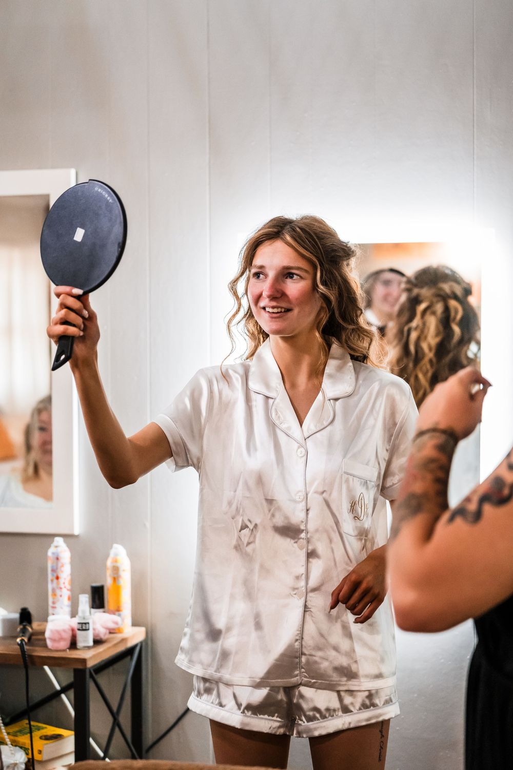 Person in white silk pajamas holding a mirror while getting ready in a dressing room.