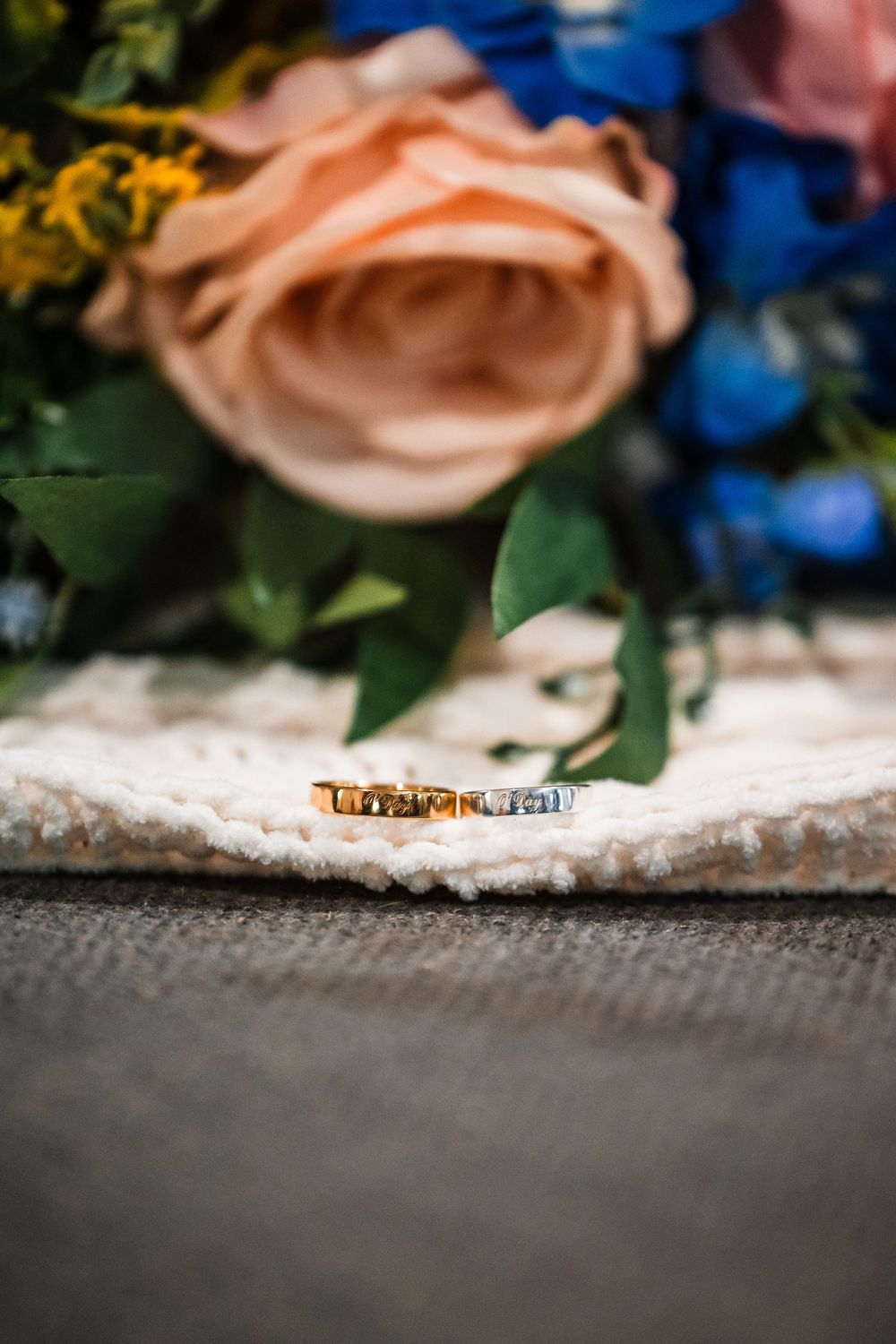 A gold wedding ring displayed on a textured surface with a blurred peach rose and blue flowers in the background.