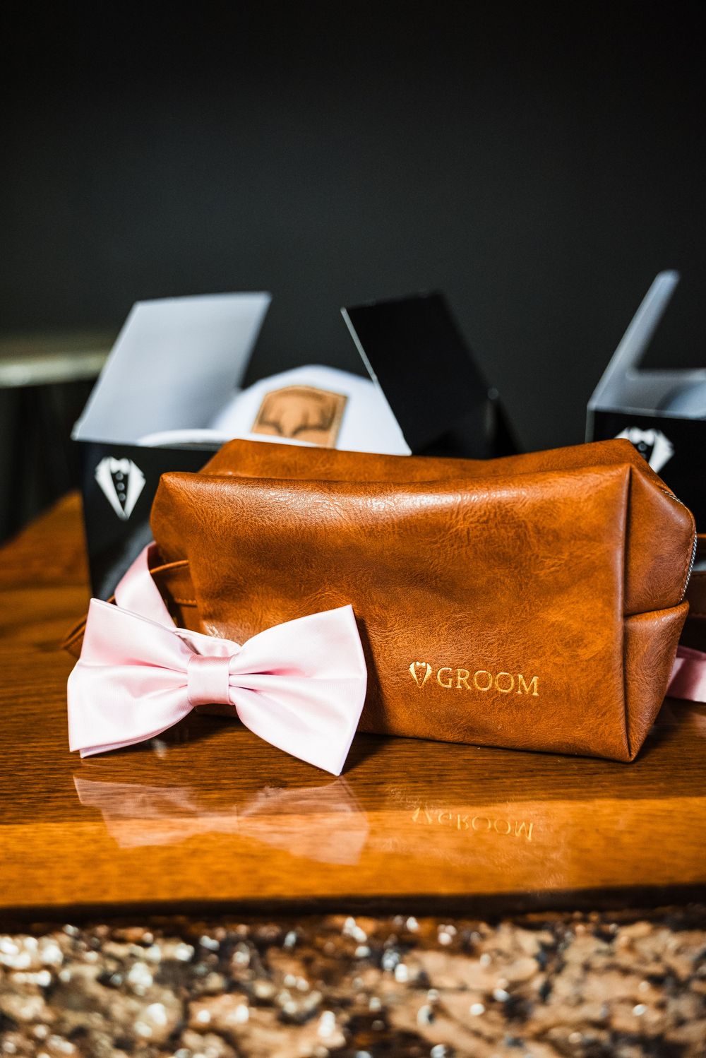 A brown leather groom's toiletry bag with a light pink bowtie on a wooden surface.