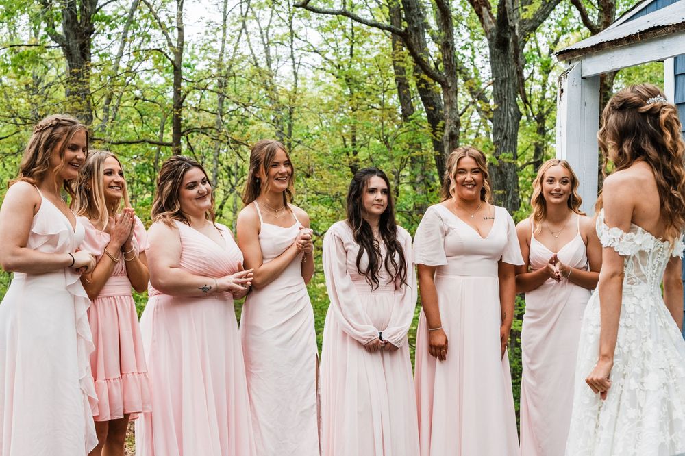 A group in soft pink and white dresses sharing a joyful moment outdoors against a natural green backdrop.