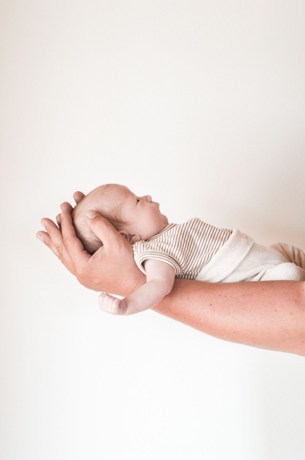 Ein neugeborenes Baby in gestreifter Kleidung ruht friedlich auf dem ausgestreckten Unterarm seines Vaters vor einem hellen Hintergrund.