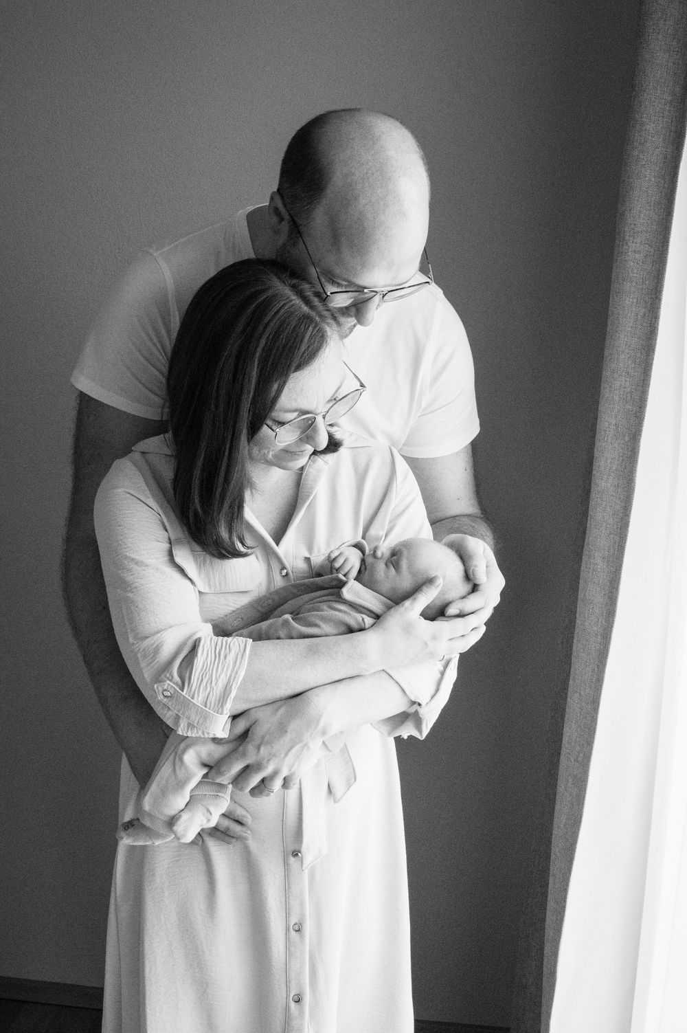 Schwarz-Weiß-Porträt von Eltern, die ihr neugeborenes Baby in dezenter Kleidung im Fensterlicht halten.