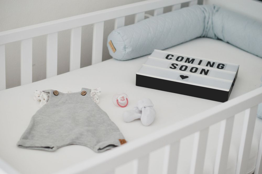 Kinderbett mit grauem Babyoverall, weißen Babysocken und einem Schild mit der Ankündigung „Coming Soon“.
