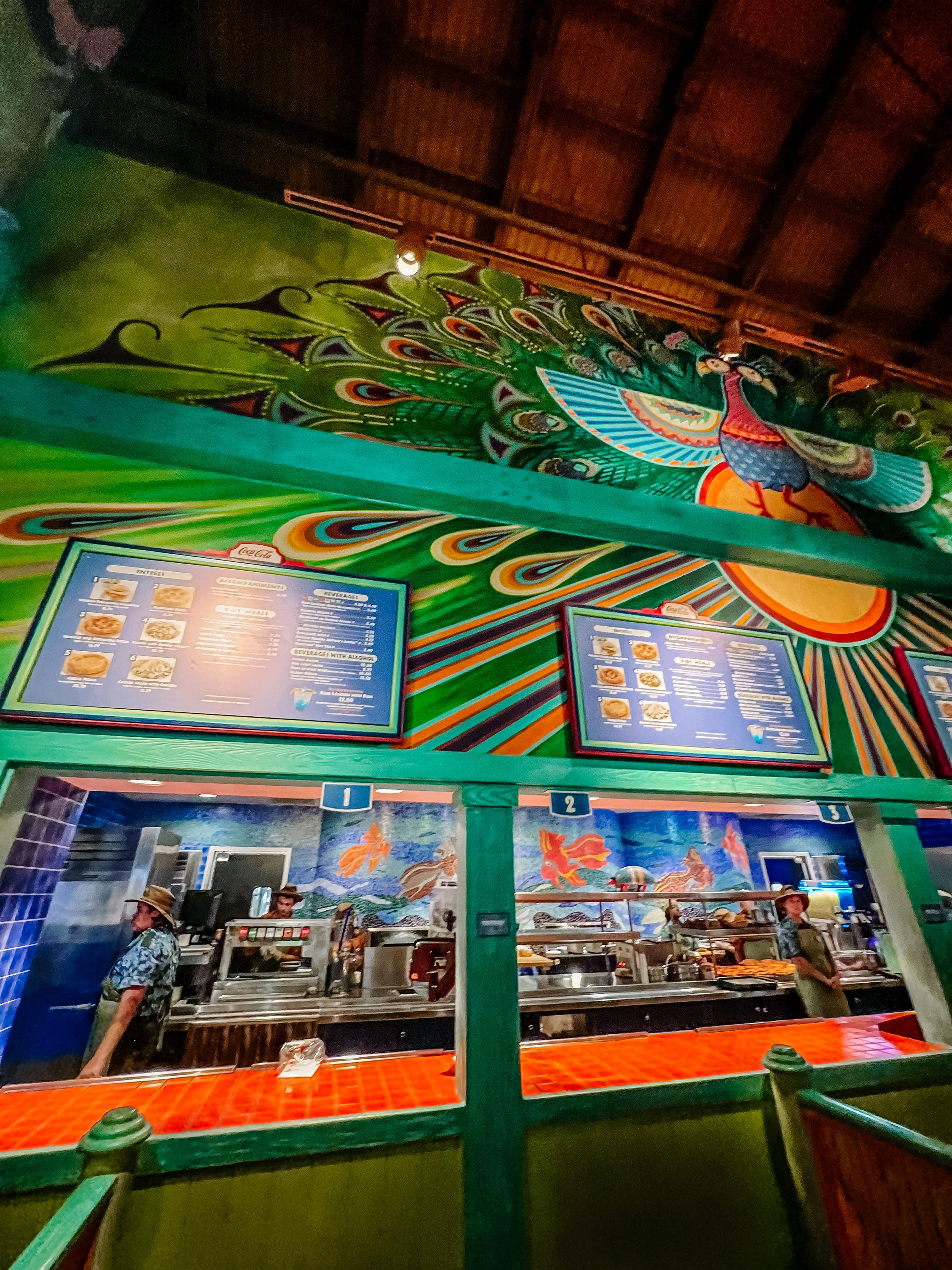 Restaurant interior with decorative peacock artwork on green walls and digital menu displays showing food items.