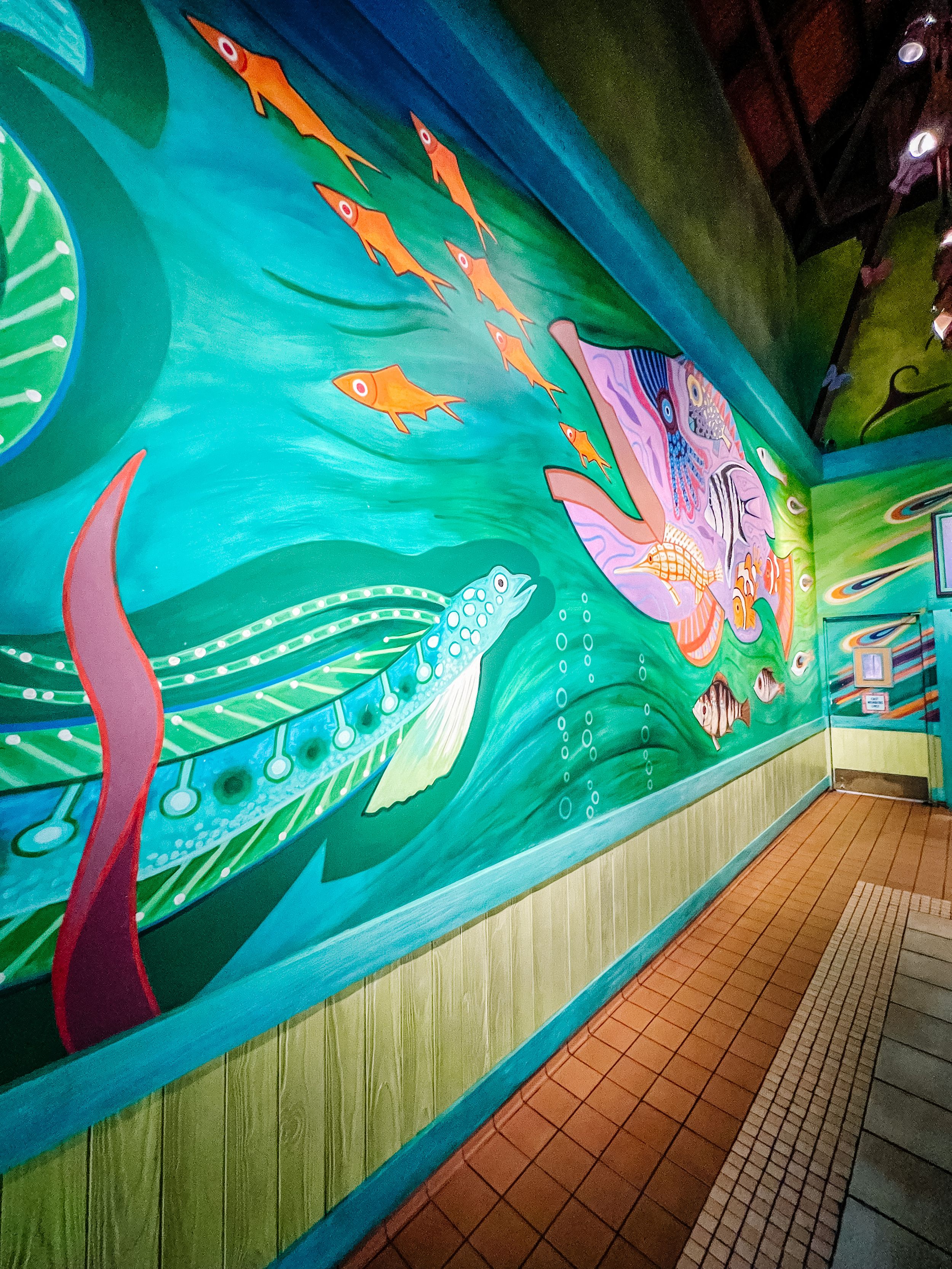 Vibrant underwater mural featuring orange fish and pink sea creatures swimming against a turquoise wall in a tiled hallway.