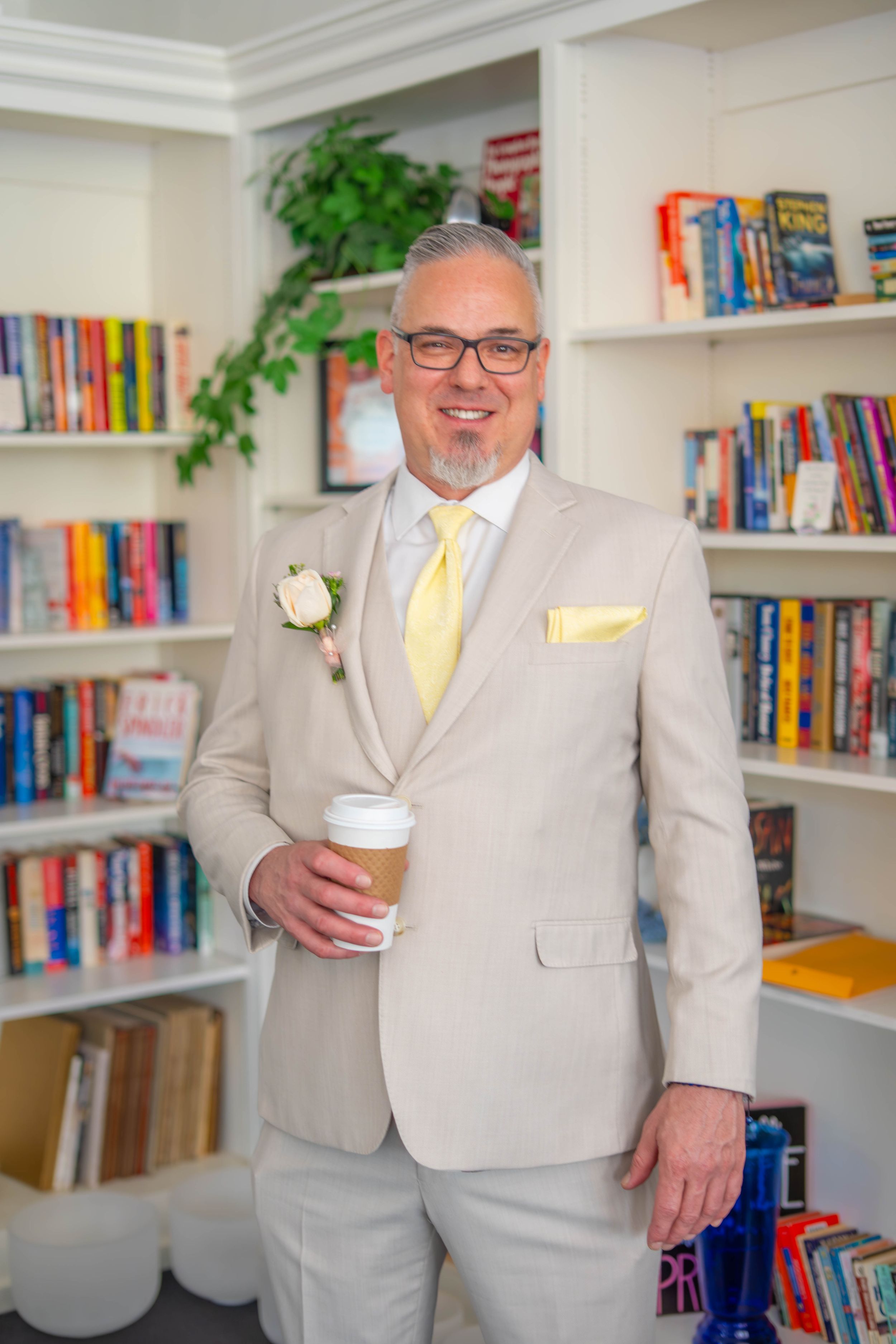 Someone wearing a light gray suit and yellow tie stands in front of bookshelves holding a coffee cup in an office setting. Quad Cities