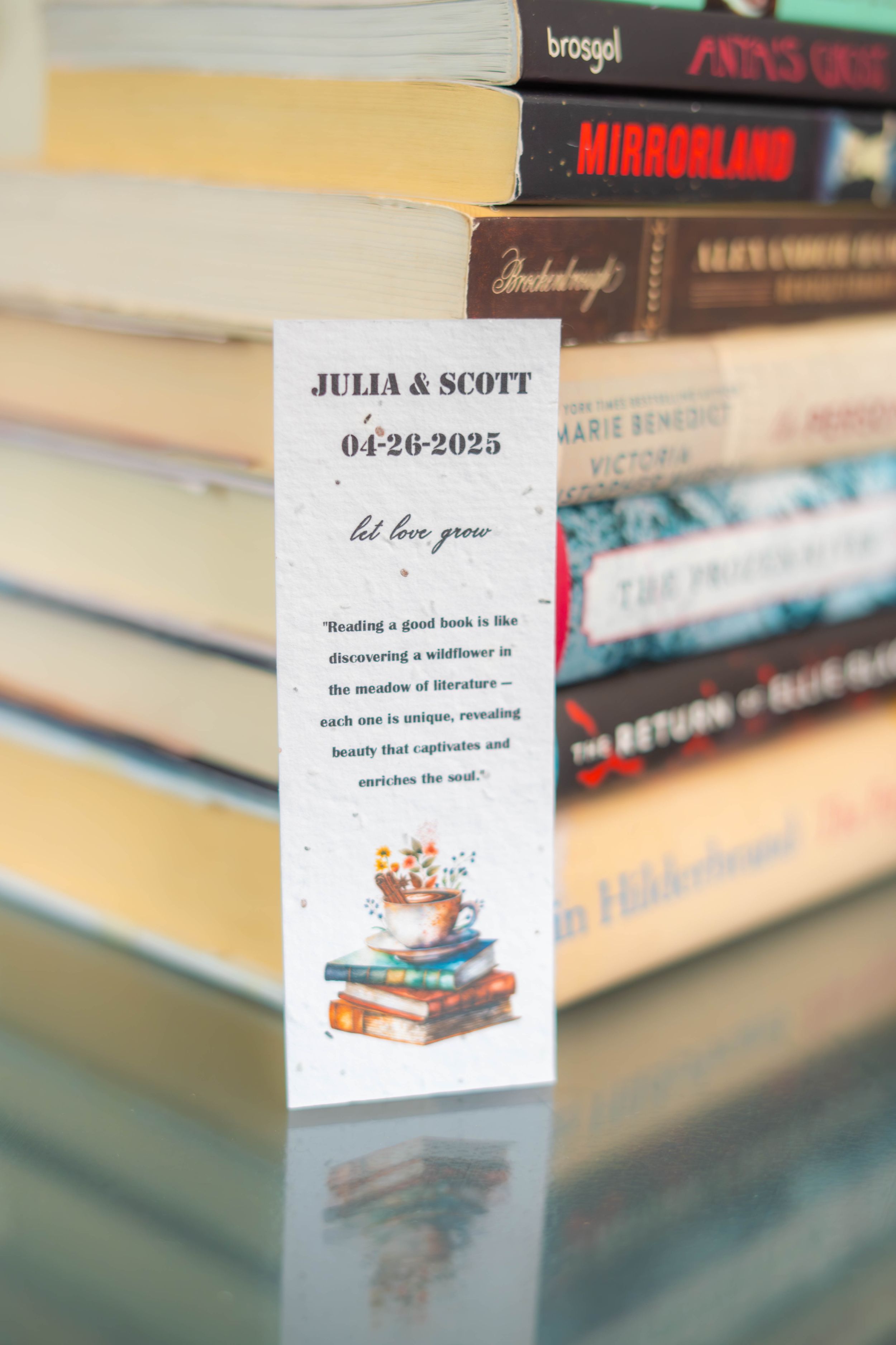 A bookmark with a wedding date and illustration of books rests against a stack of hardcover novels on a reflective surface. Quad Cities