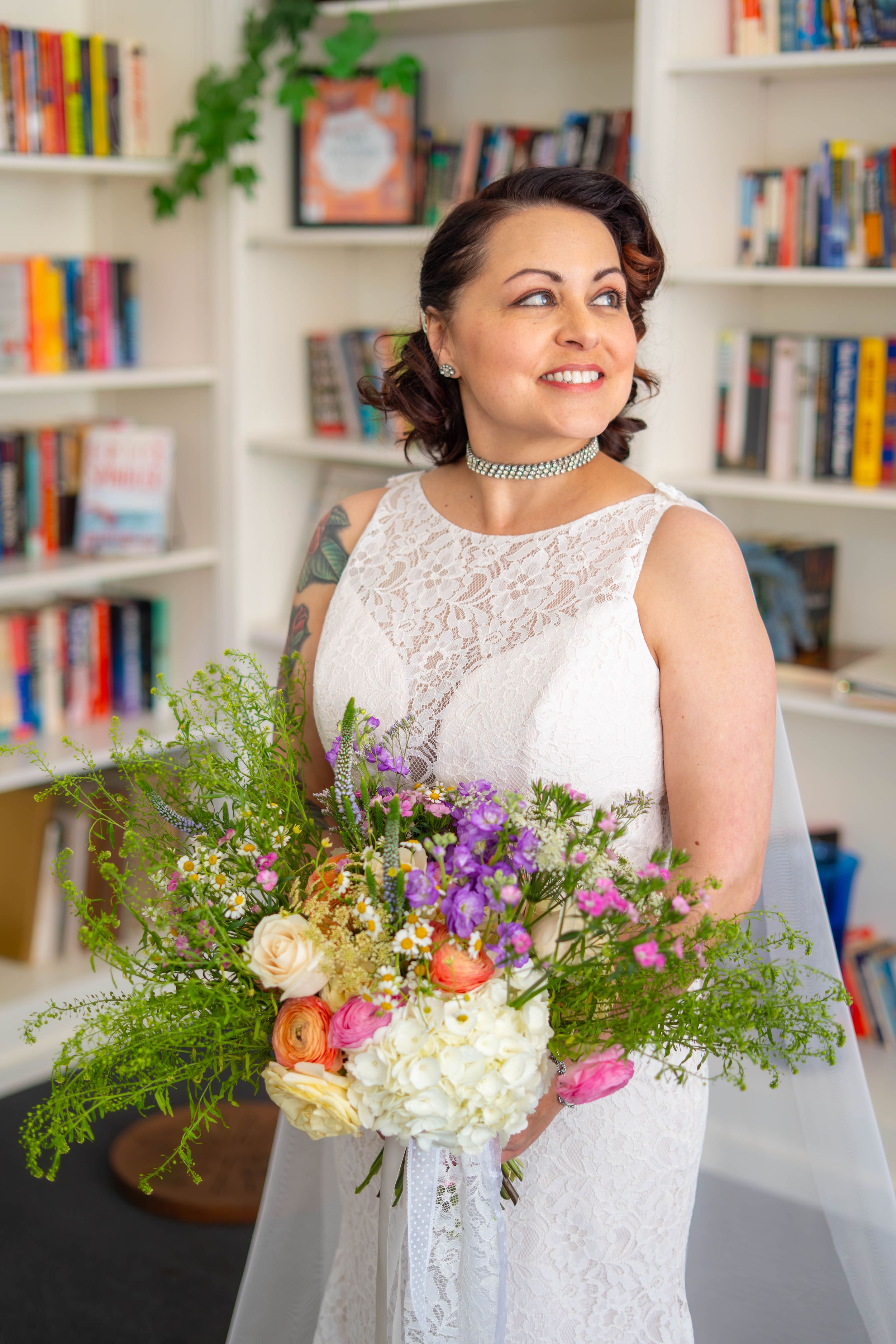 A beautiful bridal portrait showing a lace white dress and colorful wildflower bouquet against bookshelves in a library setting. Quad Cities