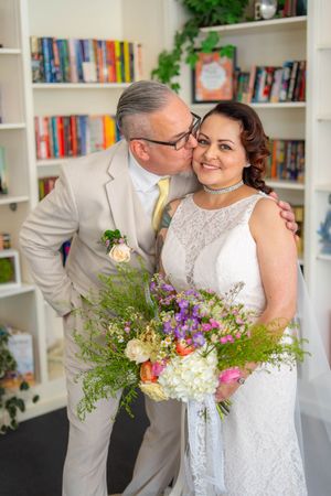 Wedding couple embraces in front of bookshelves, with the bride holding a vibrant wildflower bouquet. Quad Cities Wedding Photography