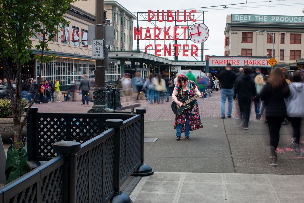 Pedestrians walking past iconic Public Market Center sign in downtown area with a guitarist perfectly still as they pass by.