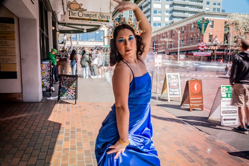 Person in blue formal dress posing dramatically outside urban storefront as crowd is motion blurred in the background