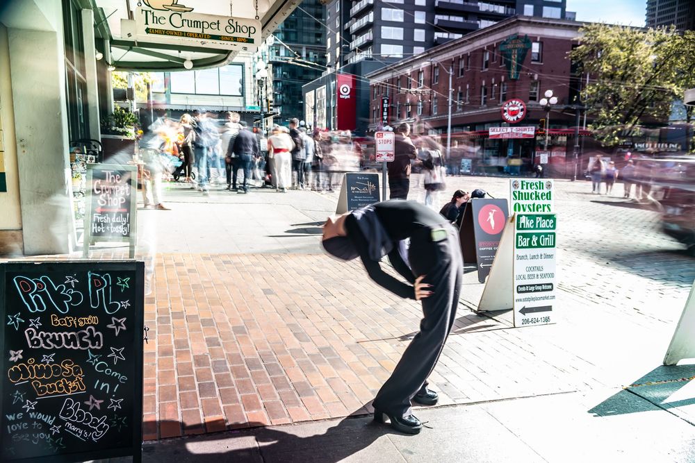 Person bending backwards gracefully in front of motion blurred crowd on brick sidewalk near outdoor market in urban setting.