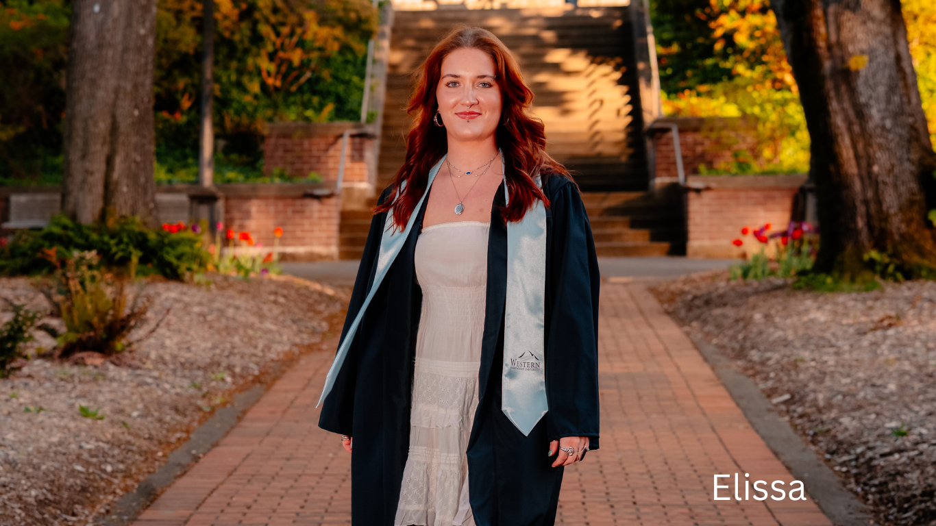 Graduate in black cap and gown with white stole standing on brick pathway during sunset.