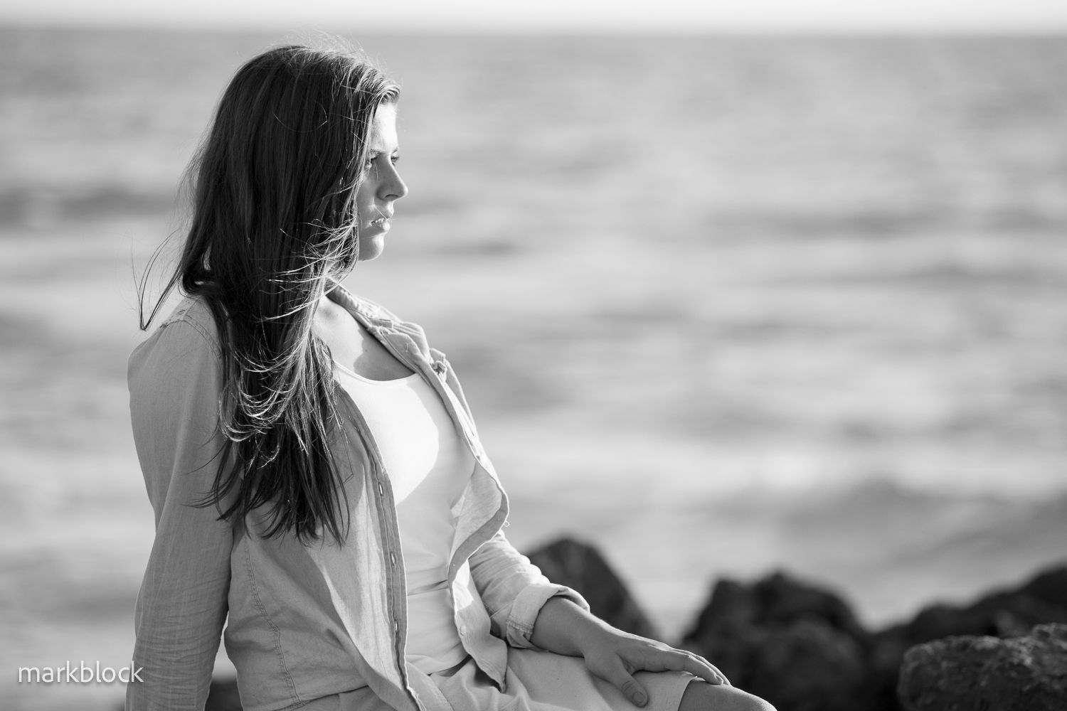A dreamy black and white portrait of someone sitting on coastal rocks with long flowing hair in the wind.