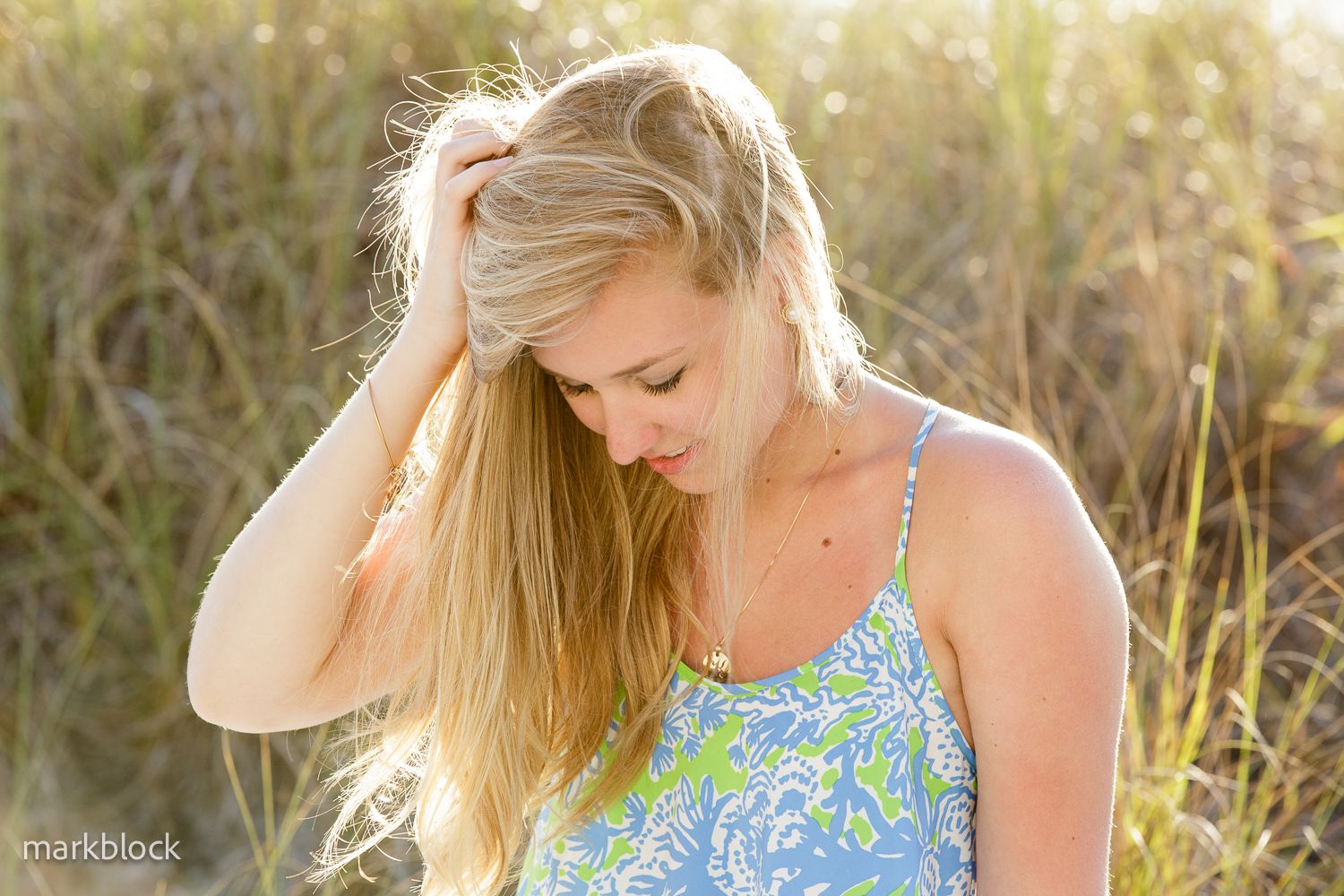 A woman in a blue floral tank top stands in a sunlit field of tall grass at golden hour.