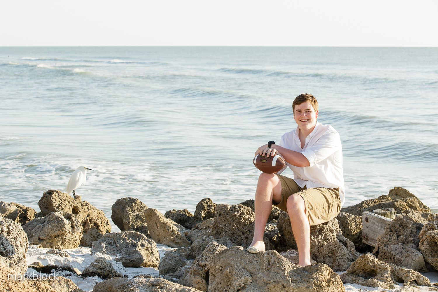 Person relaxing on coastal rocks by the ocean during sunset while wearing white shirt and khaki shorts.