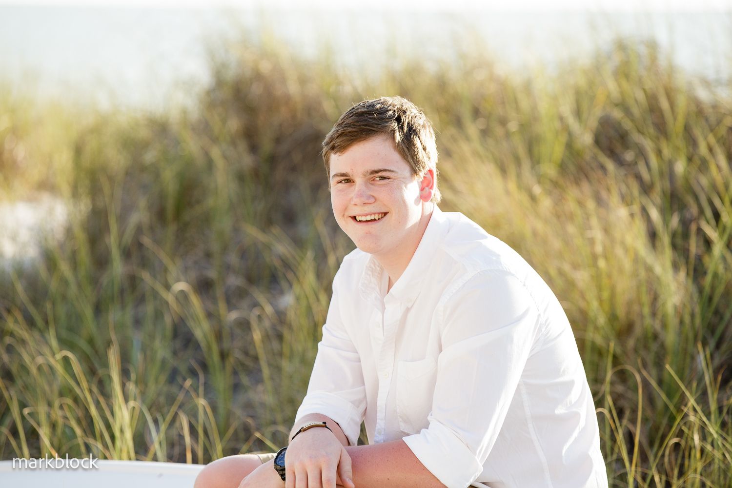 A person in a white button-down shirt sits smiling in tall grassy dunes during golden hour.