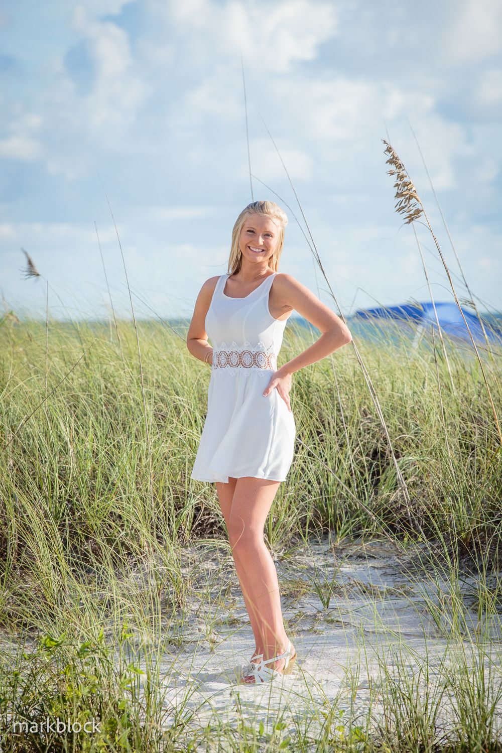 A summery portrait in a white sundress standing among beach grass and sand dunes on a sunny day.