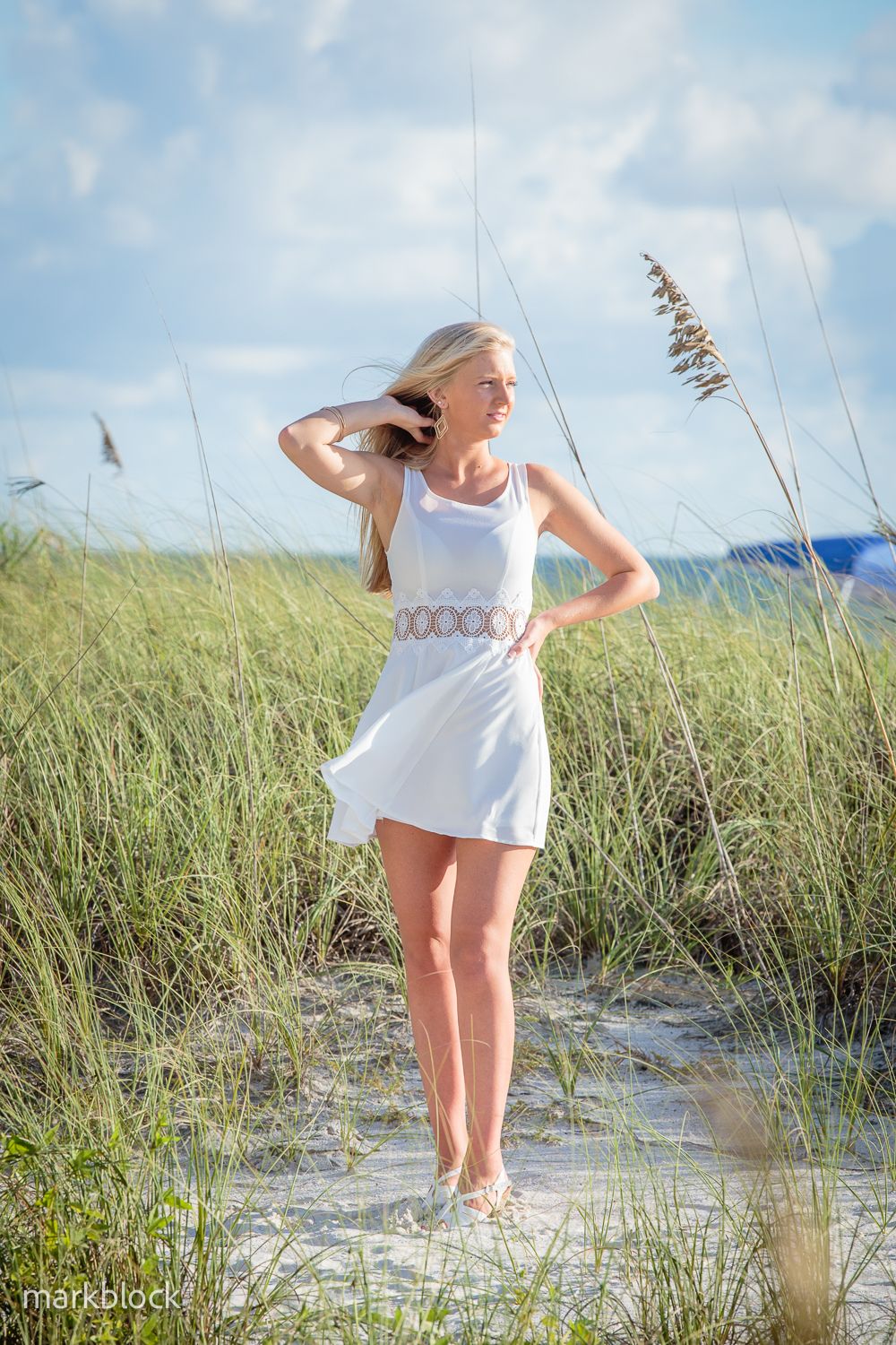 Someone in a white summer dress poses among tall beach grass on sand dunes with blue sky in the background.