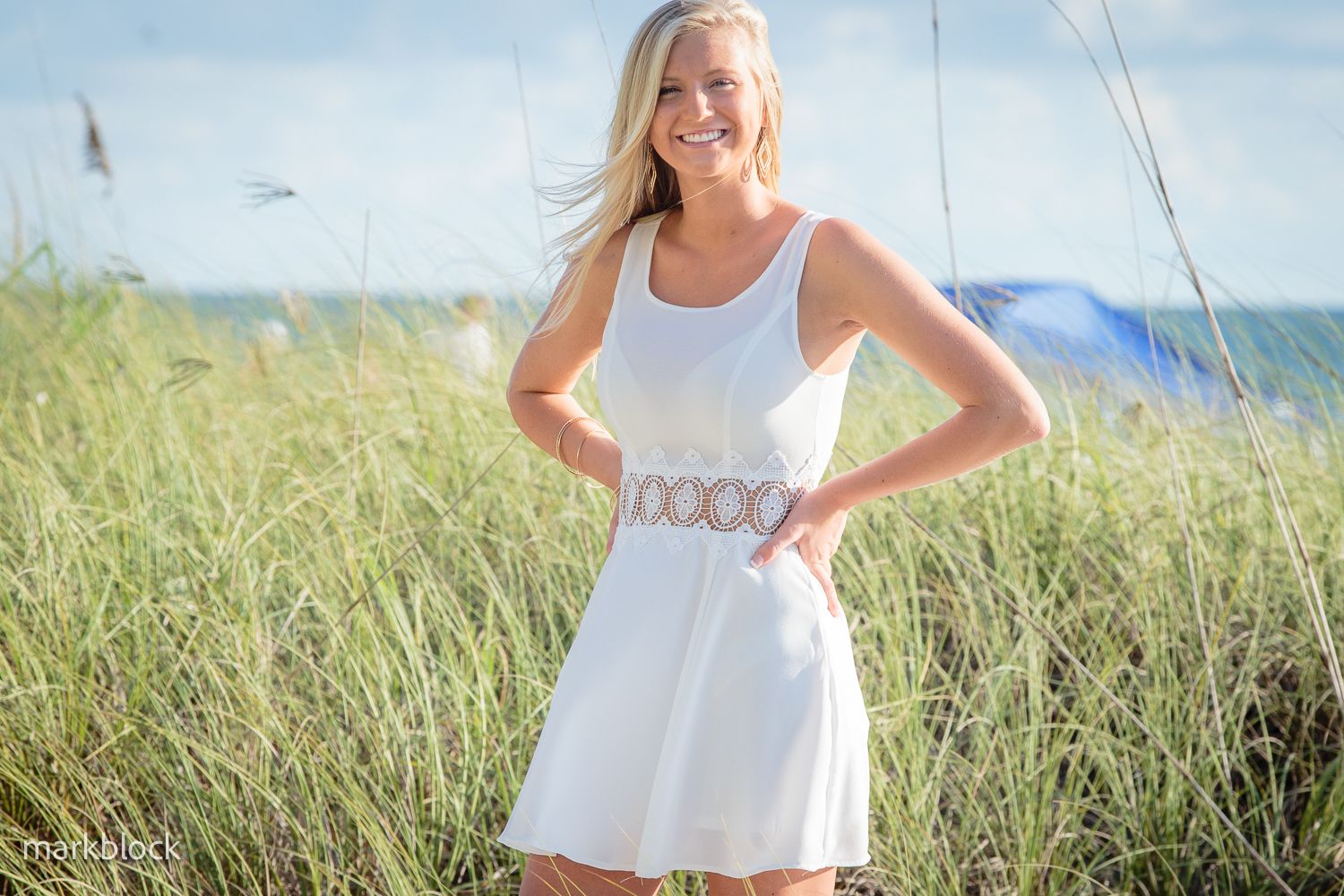 A person in a white summer dress stands smiling in tall beach grass on a sunny day.
