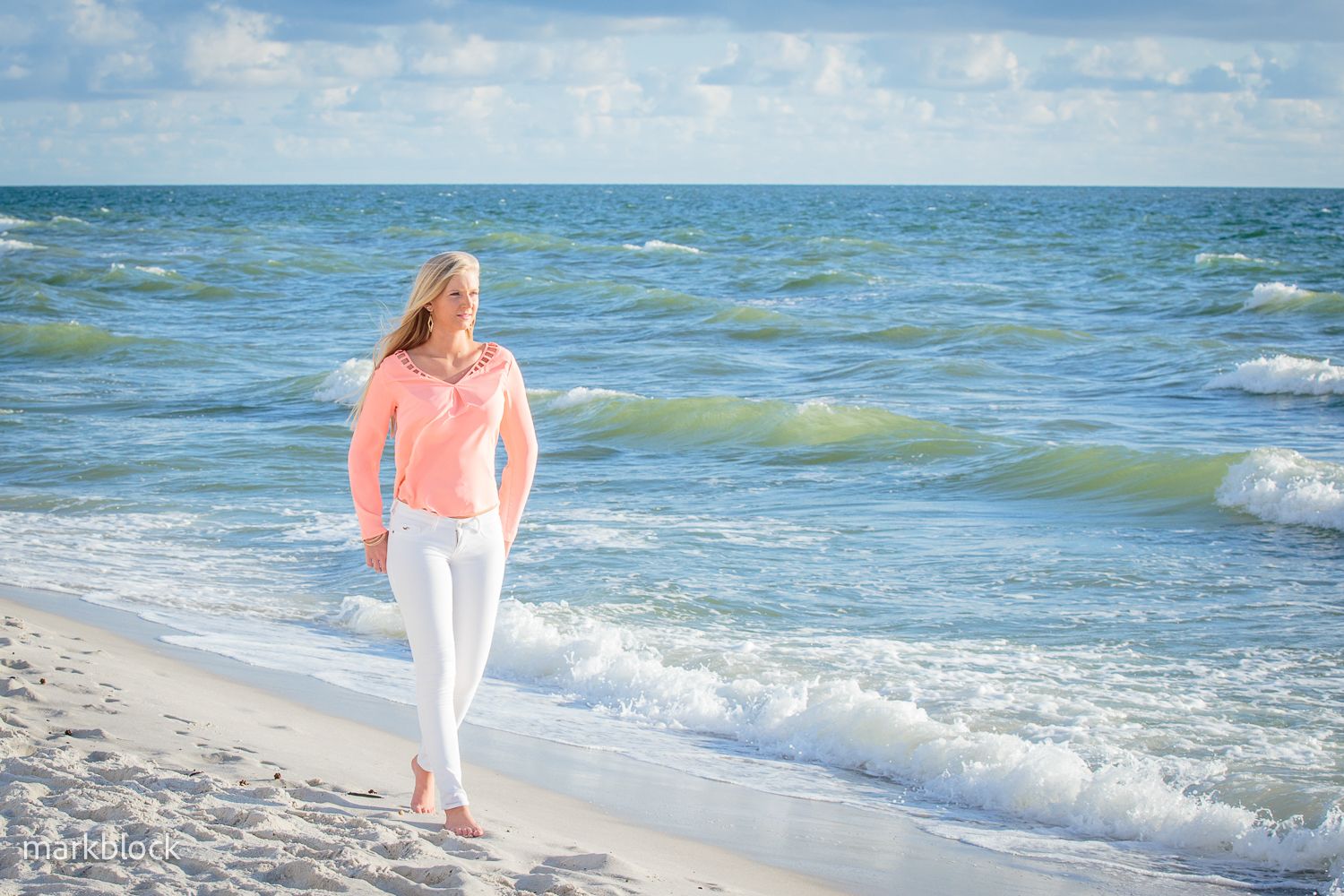 A person in a coral sweater and white pants walks along the shoreline on a sunny beach with turquoise waves.