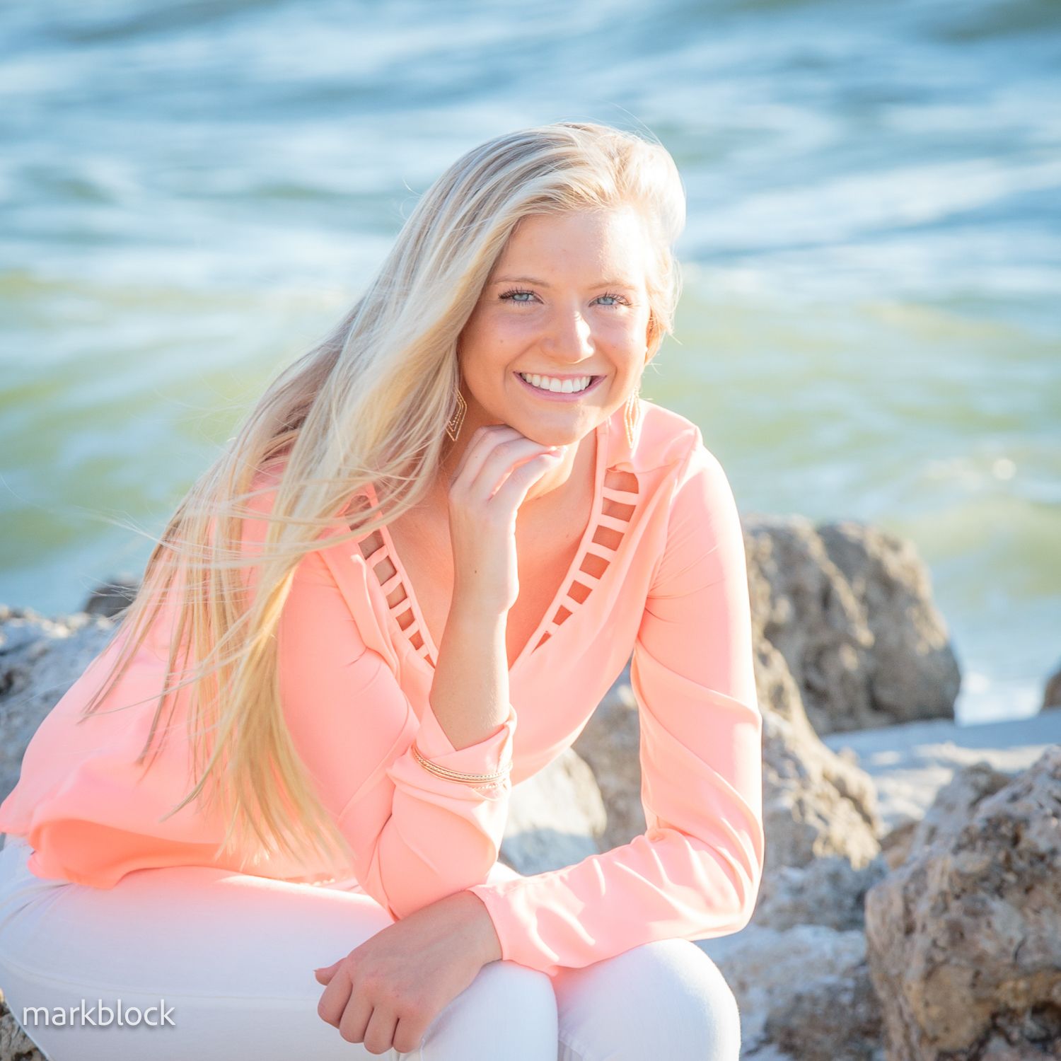 Someone sits on coastal rocks wearing a coral sweater with decorative cutouts against a blue ocean background.