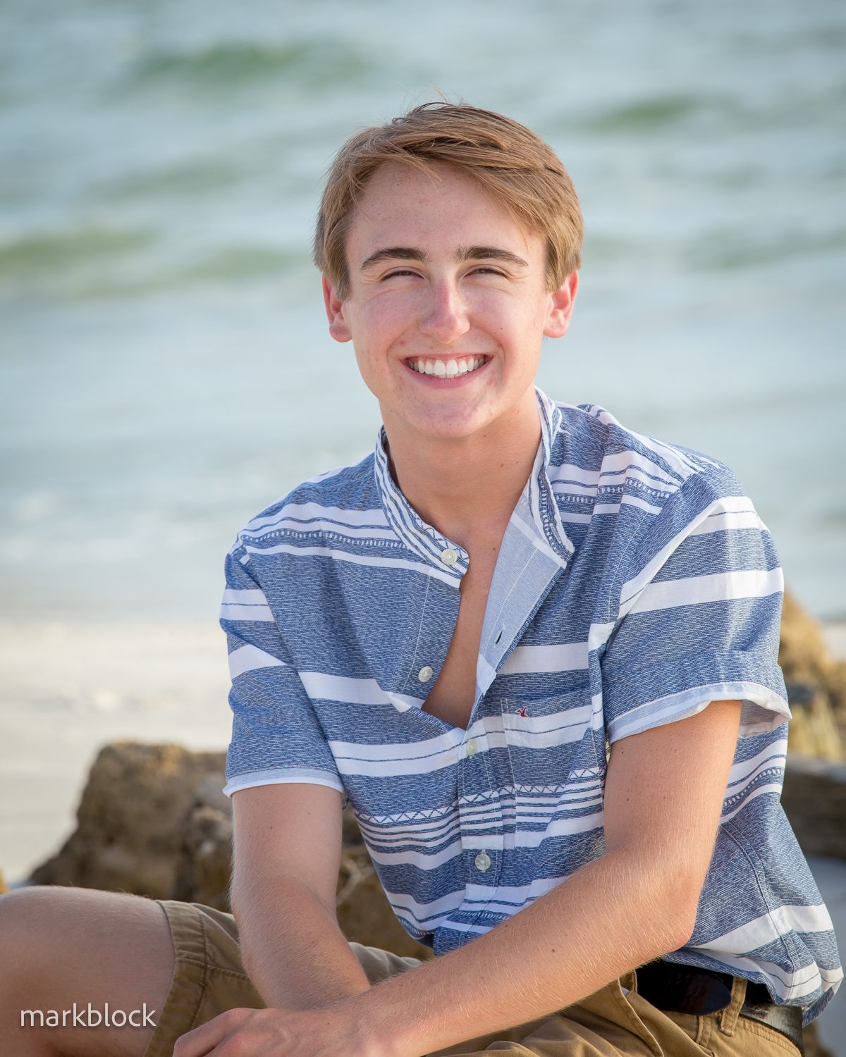 A smiling figure in a blue and white striped short sleeve shirt sits on a beach with blurred ocean waves in the background.