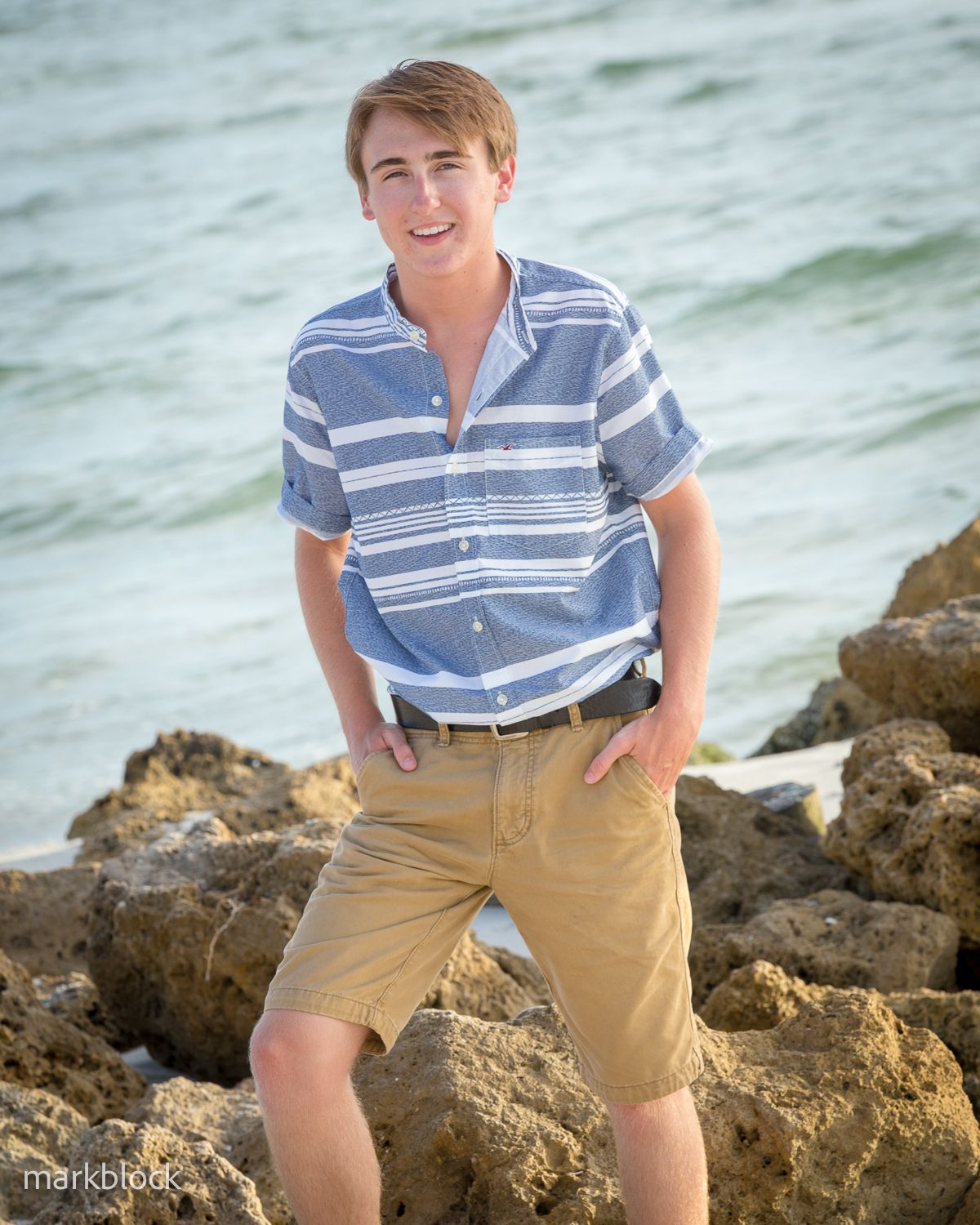 Someone wearing a blue and white striped shirt and khaki shorts poses casually on coastal rocks by the ocean.