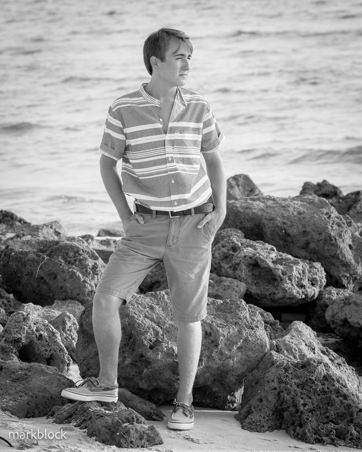 Black and white photo of someone in a striped polo shirt and shorts standing on rocky coastal terrain by the ocean.