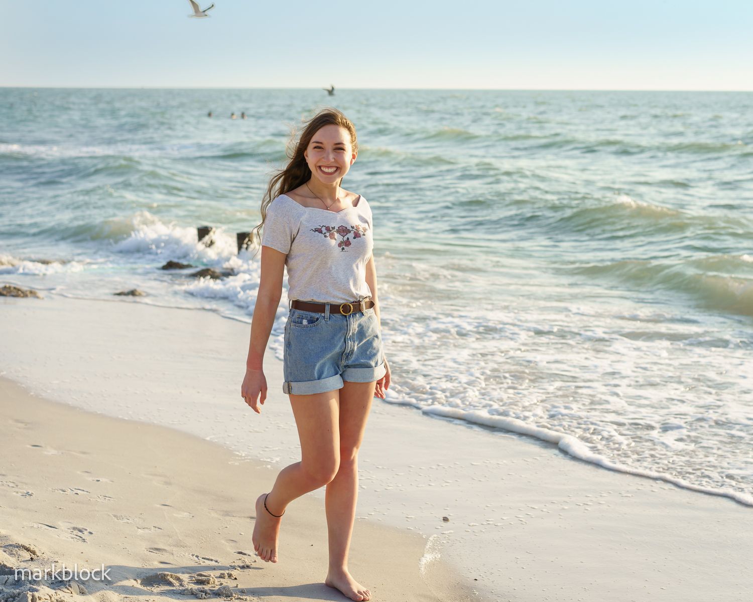 Someone walks barefoot along a sandy beach shoreline on a sunny day with waves and seagulls in the background.