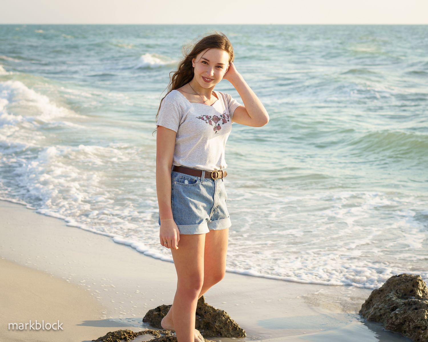 A woman in a white t-shirt and denim shorts stands on a rocky beach with turquoise waves crashing behind her.