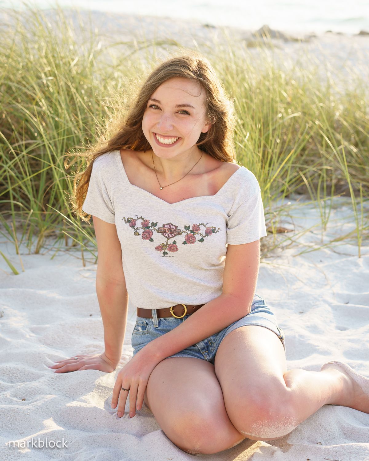 Person sitting on white sandy beach with tall beach grass in background on sunny summer day.