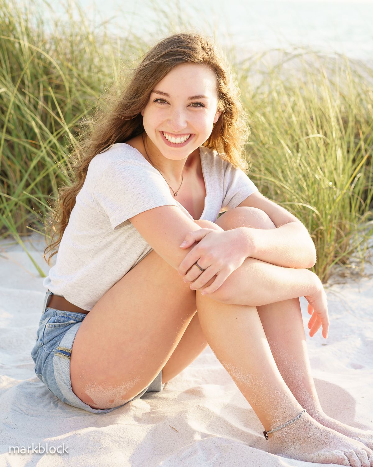 Person in white t-shirt and denim shorts sits on sandy beach surrounded by tall grass on sunny day.