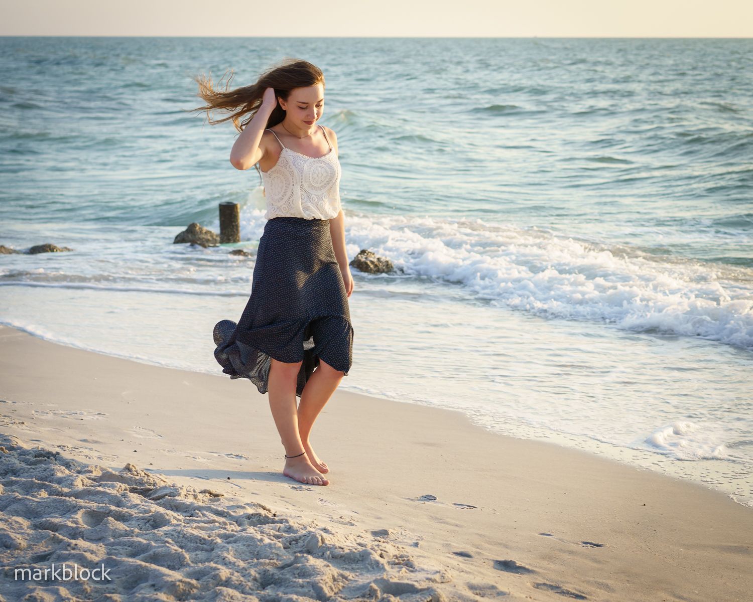 A young woman in a navy skirt walks barefoot along a sandy beach at sunset with ocean waves in the background.