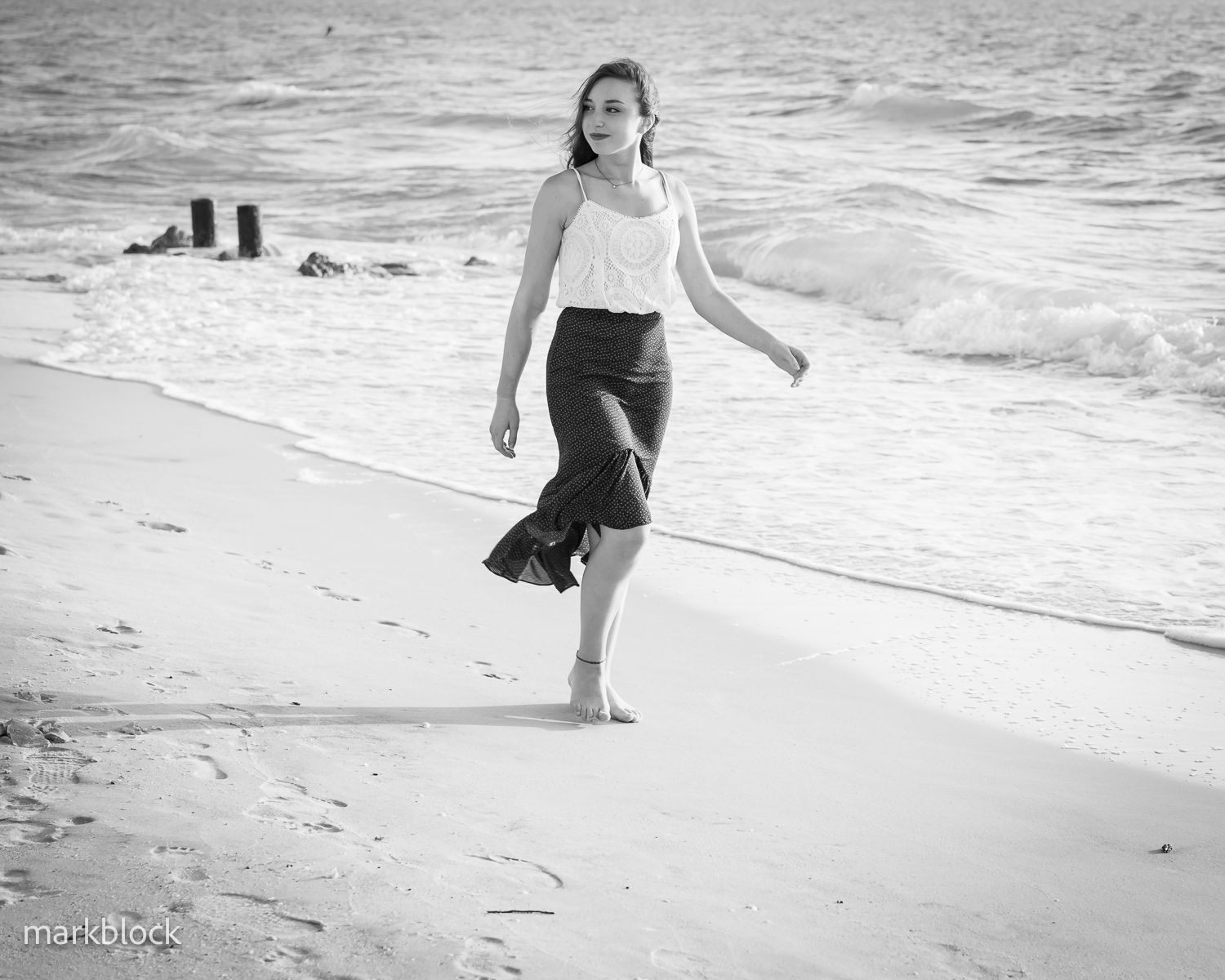 A black and white photograph shows someone walking barefoot along a beach shoreline, wearing a blouse and dark skirt.