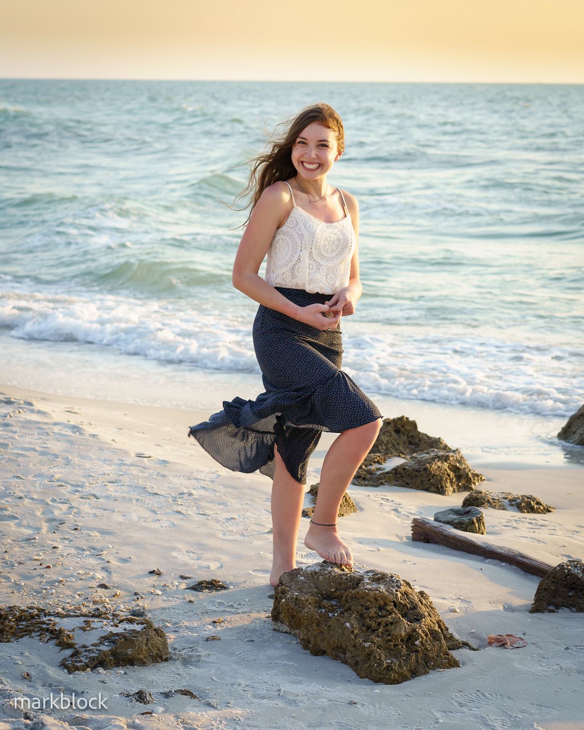 A person in a white top and navy skirt poses on a beach at sunset with waves and rocks in the background.
