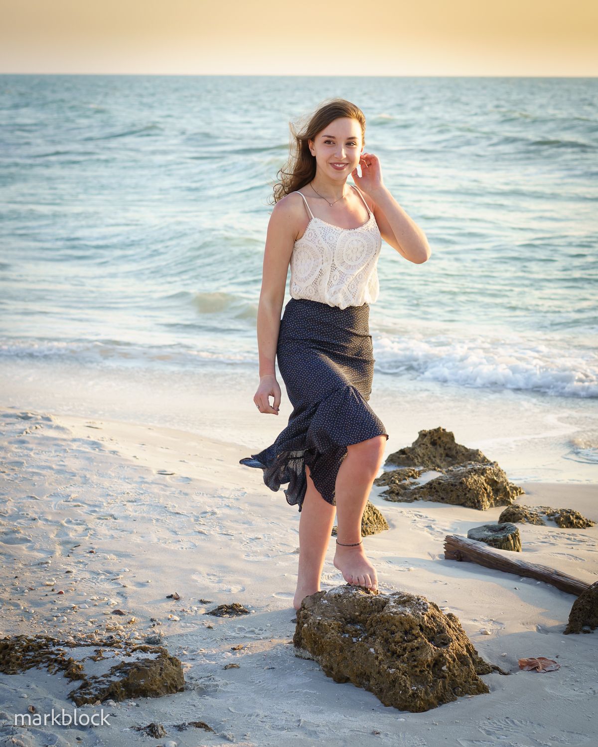 Woman in white tank top and navy skirt poses at sunset on a beach with rocks and waves in the background.