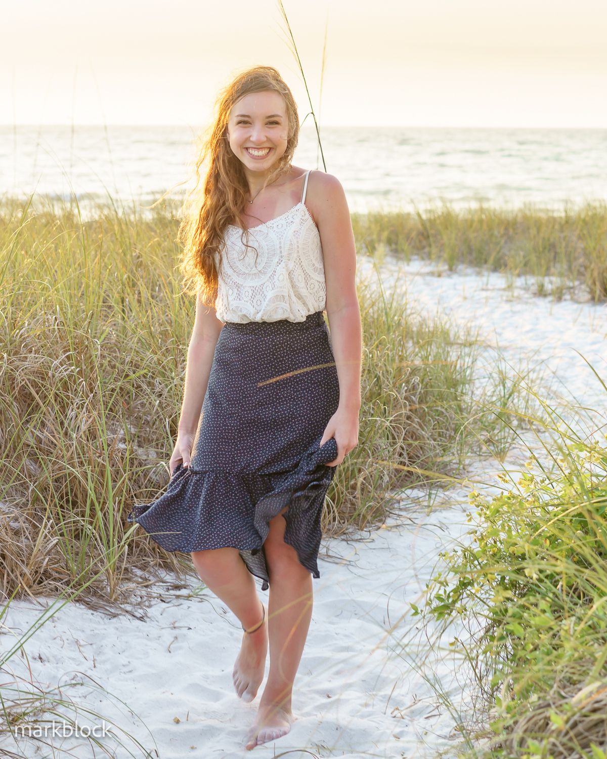 Person in white top and navy ruffled skirt walks barefoot along sandy beach path at sunset by the ocean.