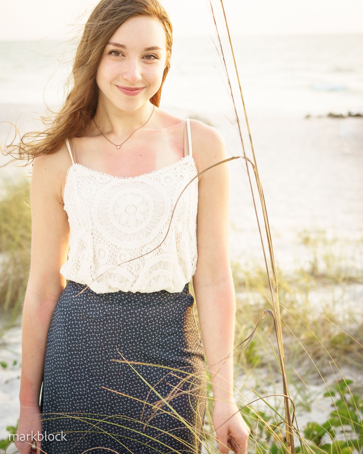 A summery coastal outfit featuring a white lace tank top and patterned navy skirt standing among beach grass at sunset.