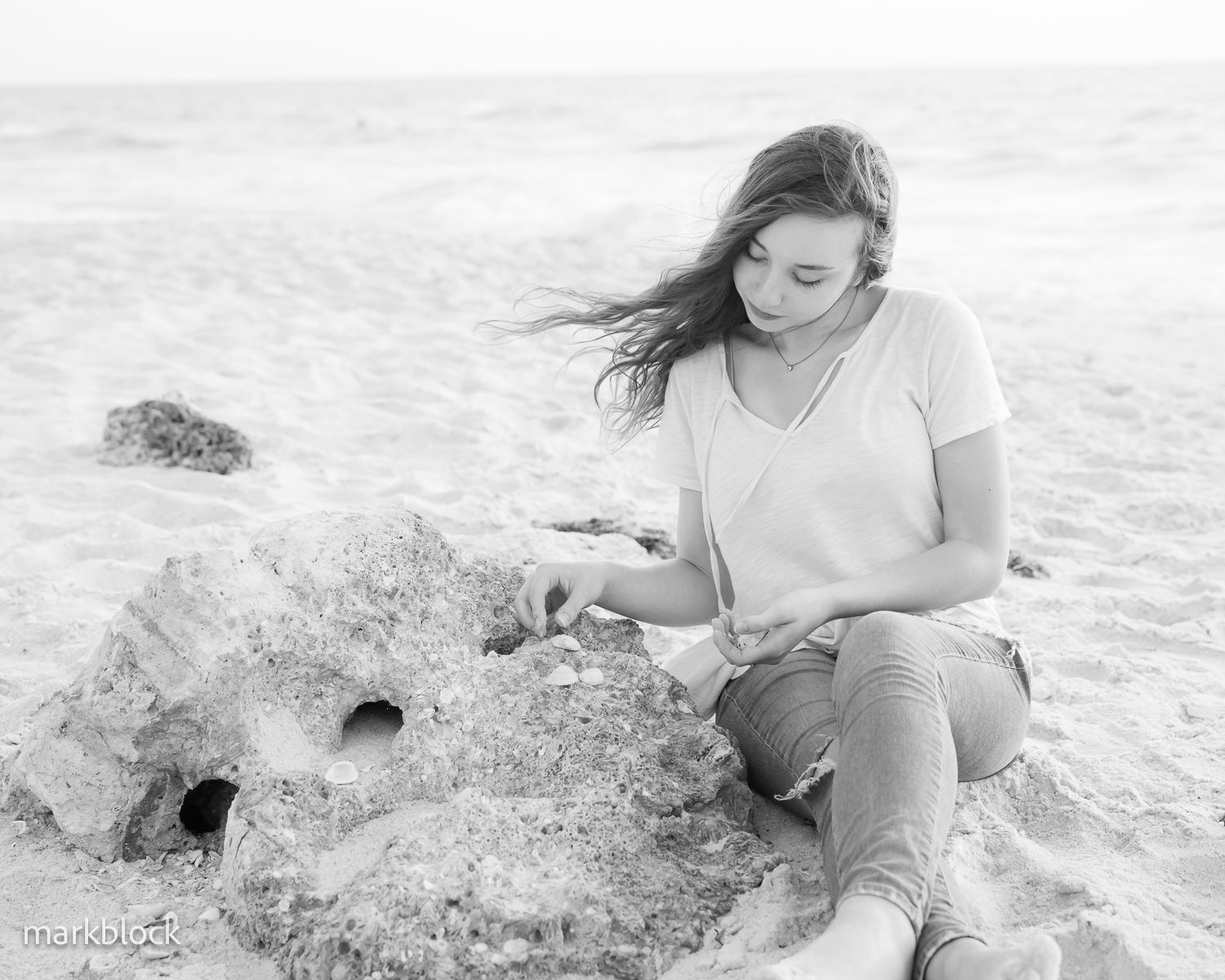 A person in casual attire sits on a sandy beach while building or sculpting something in the sand on a cloudy day.