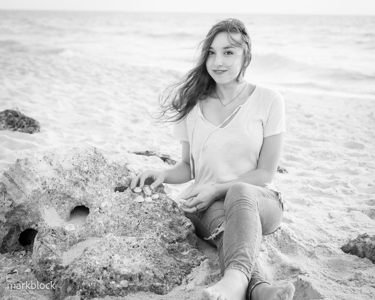 A person in a white v-neck shirt and jeans sits casually on rocky beach shores with wind-blown hair.