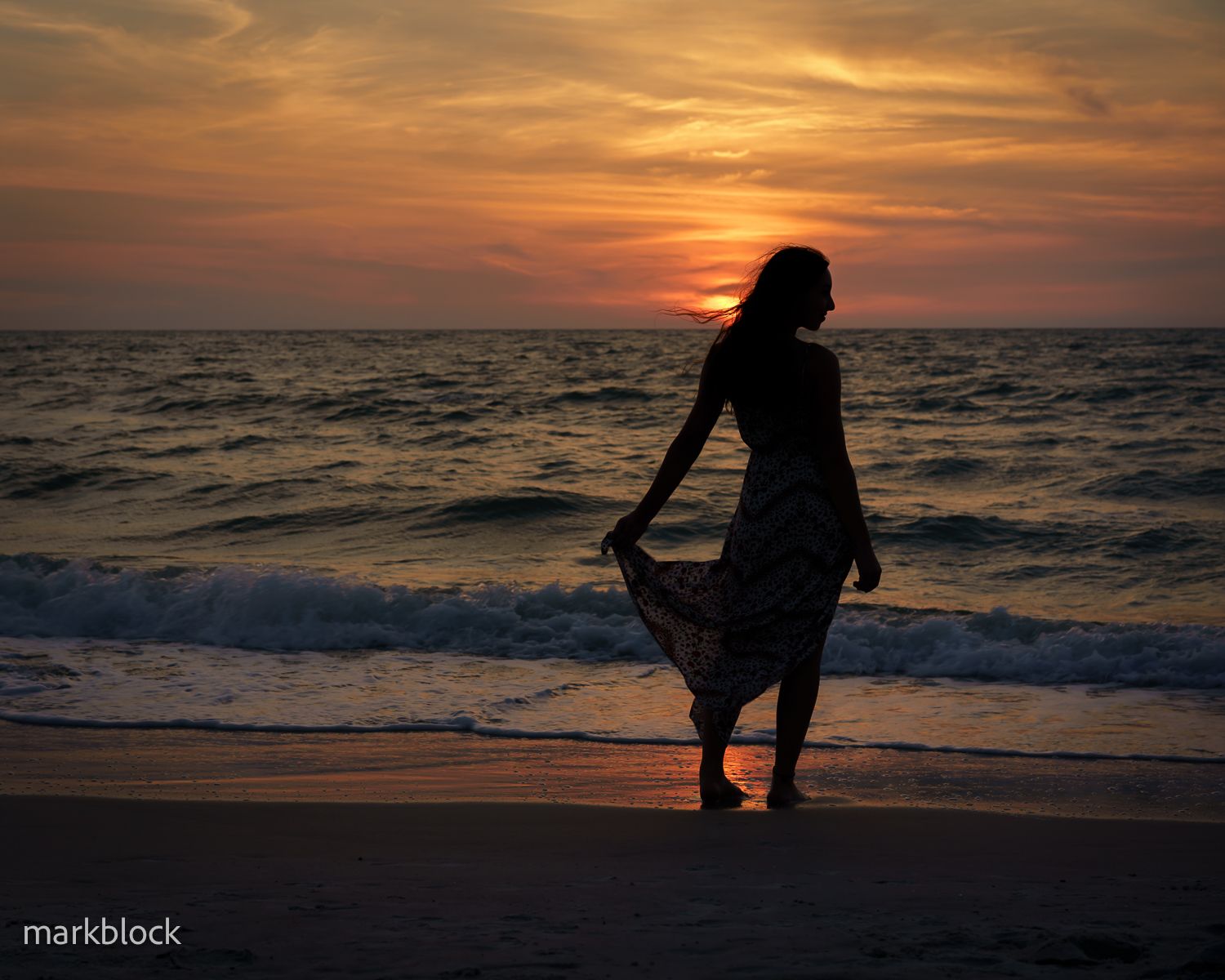 A silhouetted figure stands on the beach at sunset with waves lapping at the shore and orange clouds in the sky.