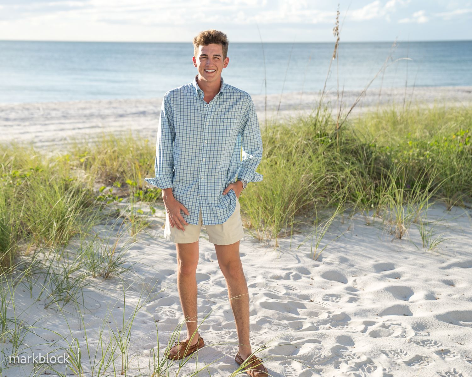 A person wearing a checkered button-down shirt and khaki shorts stands smiling on a sunny beach with grass and ocean views.