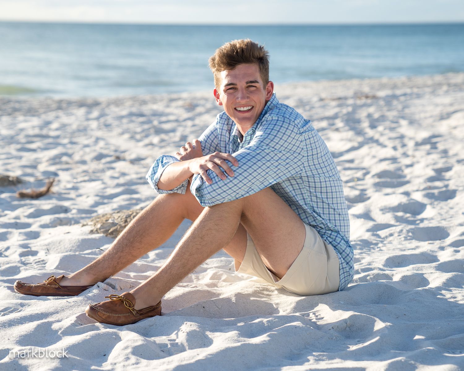A person sits on white sandy beach wearing a blue checkered shirt and khaki shorts on a sunny day.