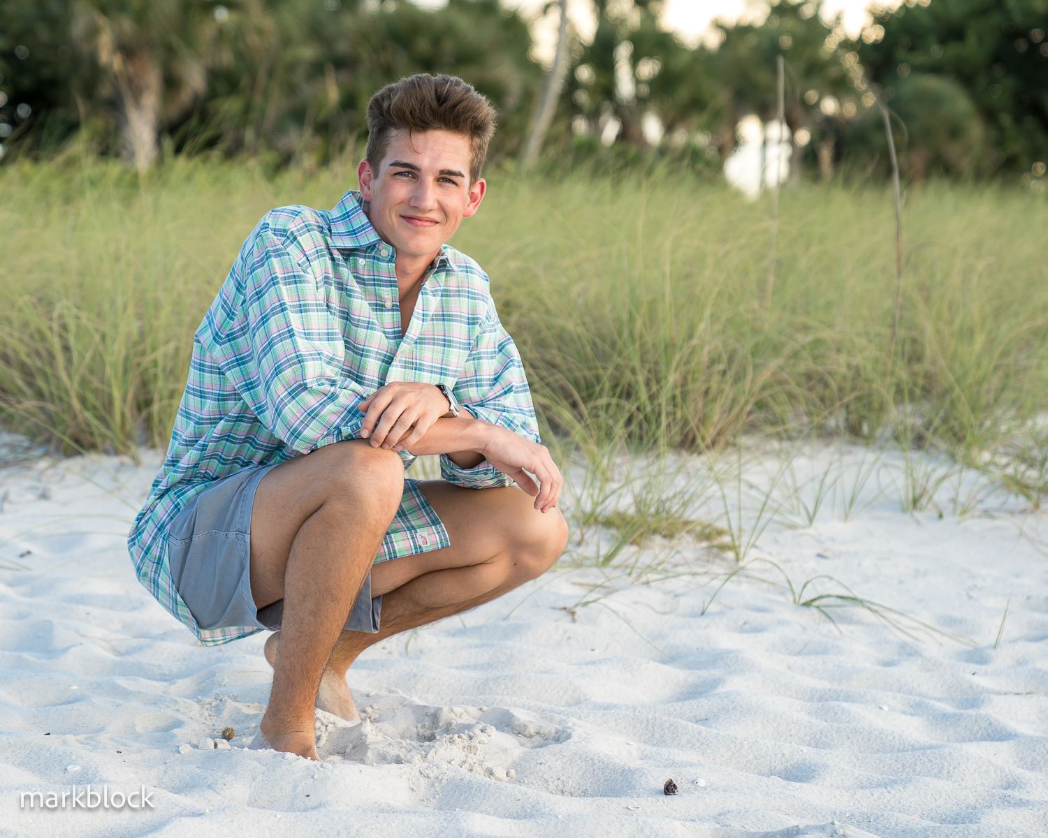 A person in blue plaid kneels on white sand with beach grass and trees in the background.