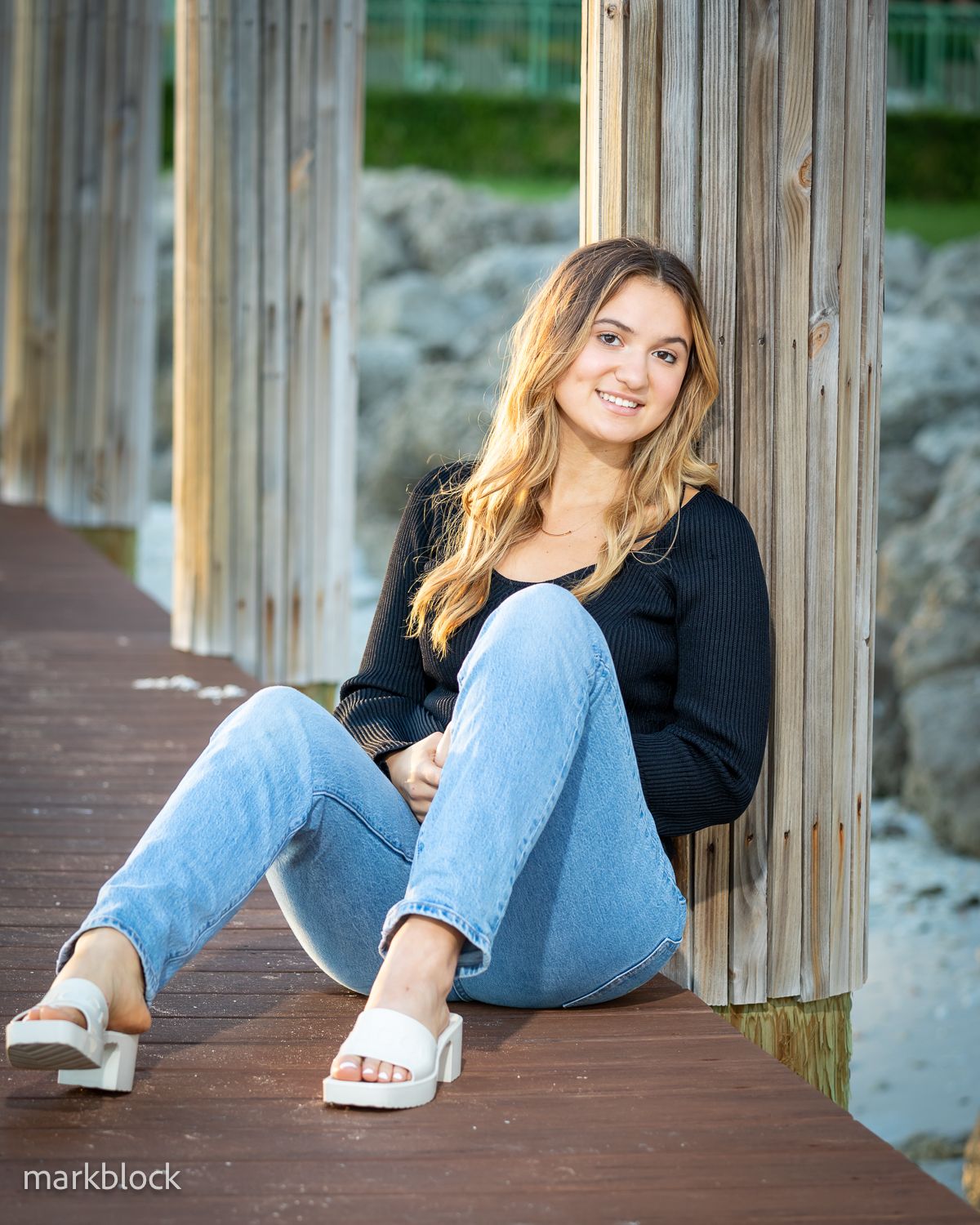 A person in a black sweater and light blue jeans sits casually on a wooden deck by the water.