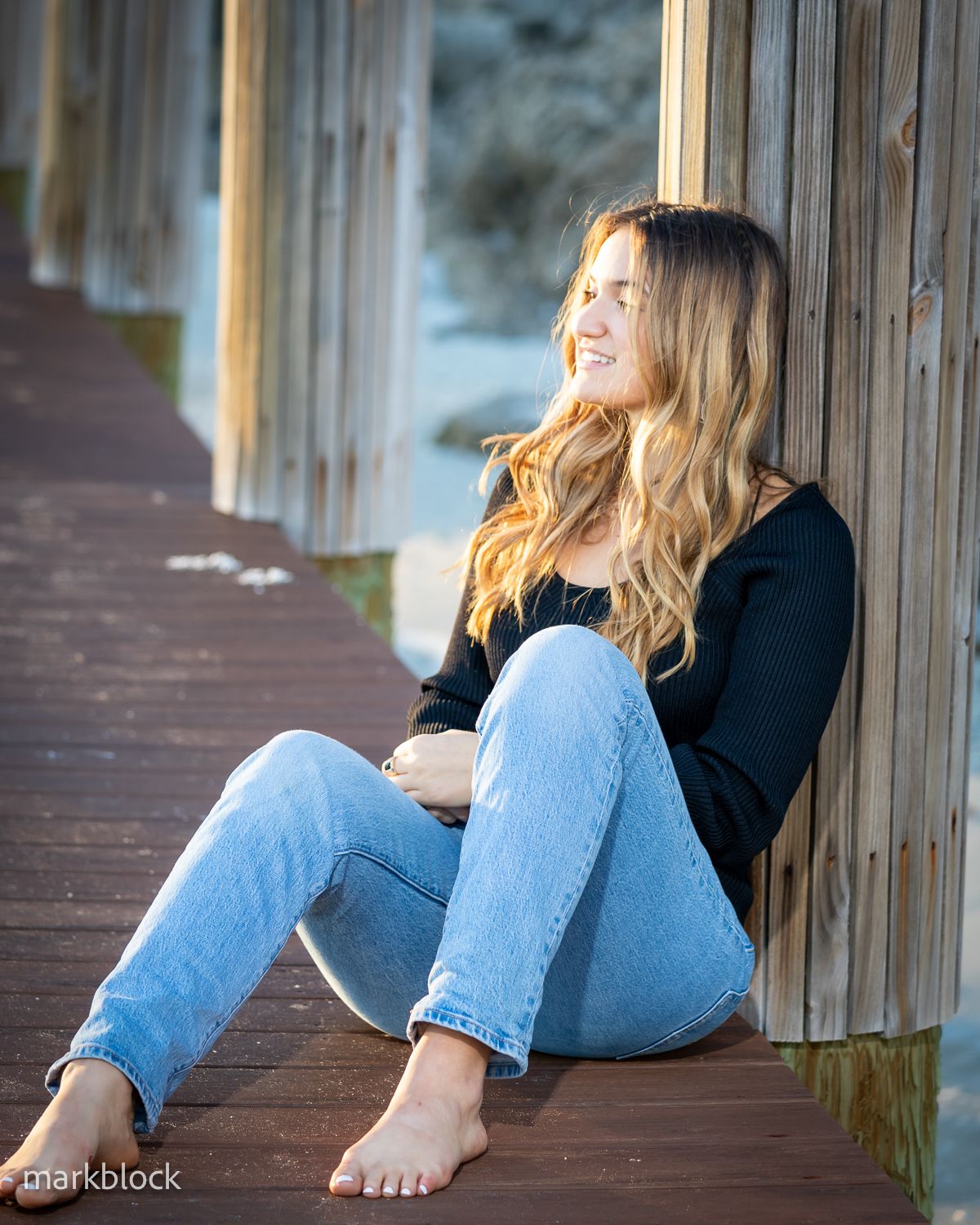 Someone with long wavy blonde hair sitting barefoot on a wooden deck wearing jeans and a black sweater.