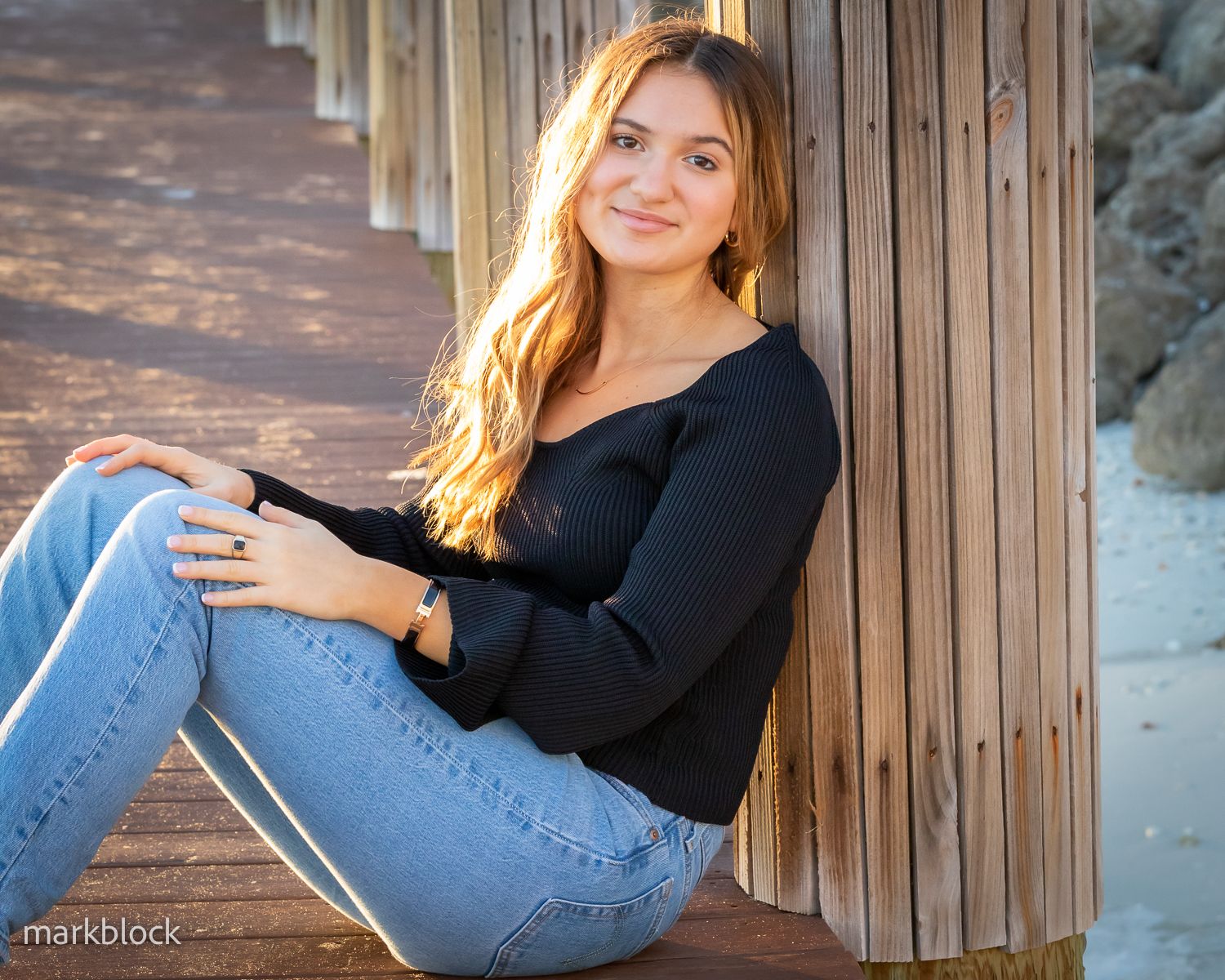 A casual outdoor portrait of someone in a black sweater and jeans sitting against wooden planks.