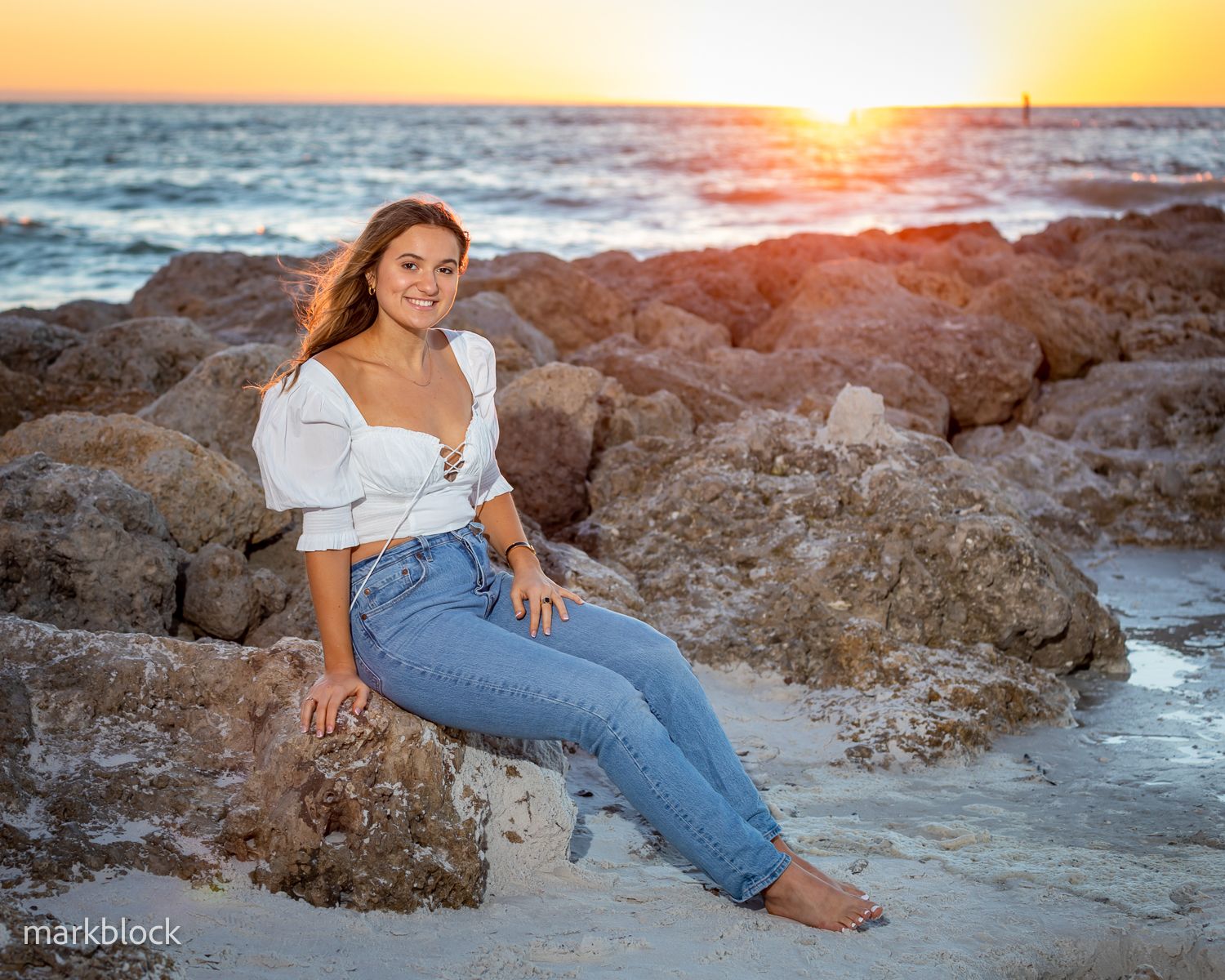 Person in white top and jeans sits on coastal rocks during golden sunset over ocean.