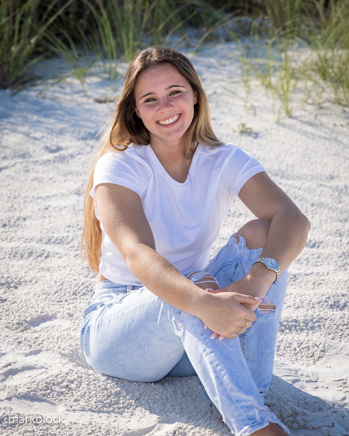 Person in casual white t-shirt and light jeans sits on sandy beach with grass in background during sunny day.